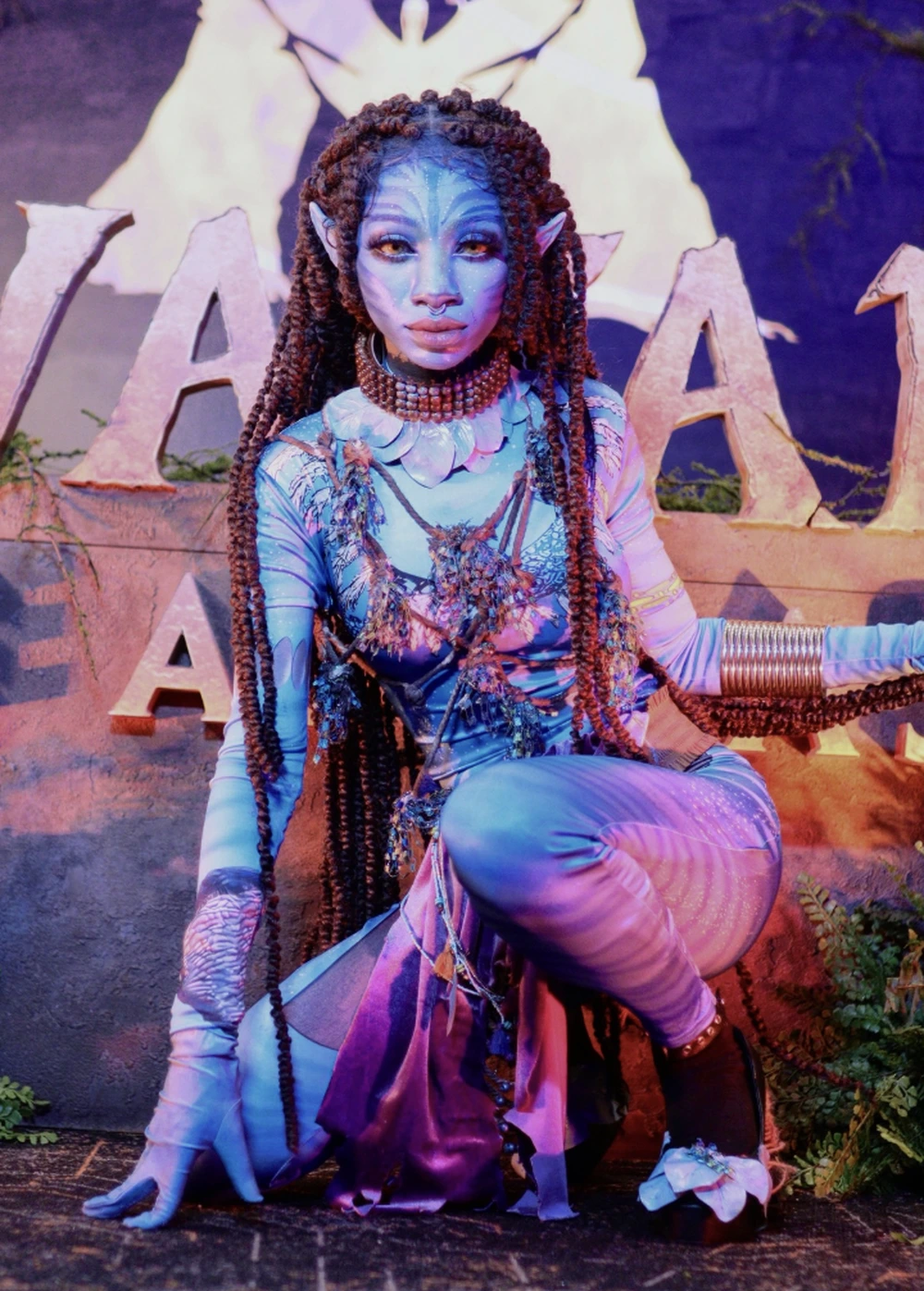 Seated blue and purple avatar costume with long braids, pointed ears, layered jewelry, and fantasy backdrop.