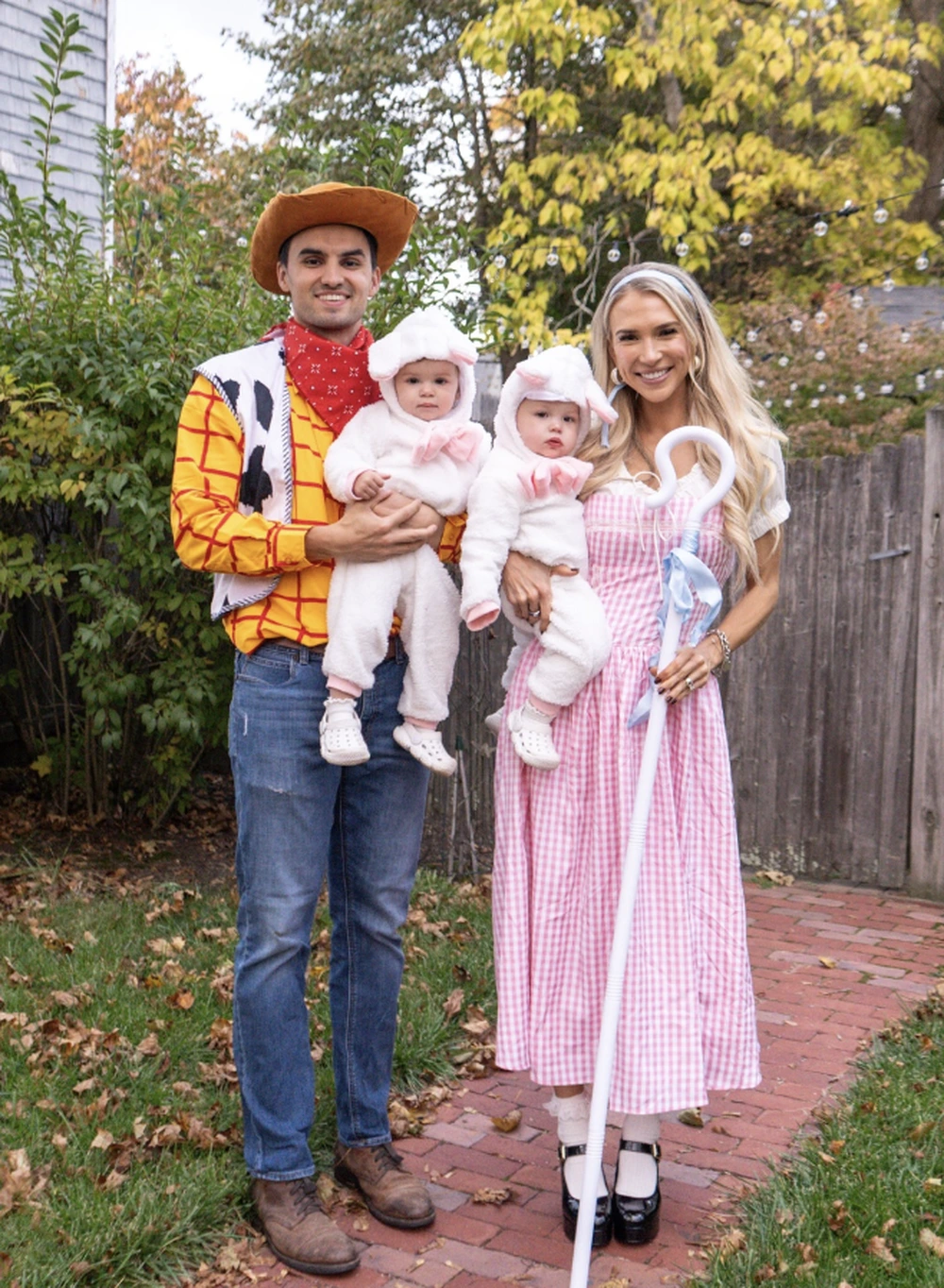 A man and woman hold twin babies while dressed as Woody and Bo Peep, standing on a brick path.