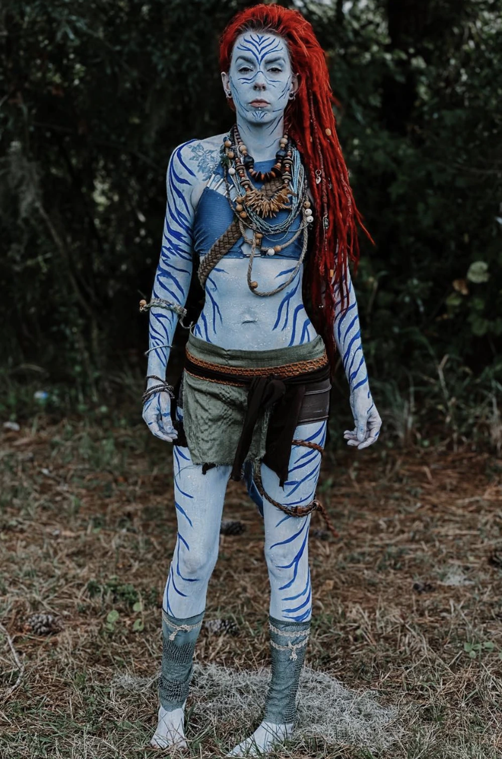 Standing blue avatar with red dreadlocks, layered necklaces, painted stripes, green wrap skirt, and woodland setting.