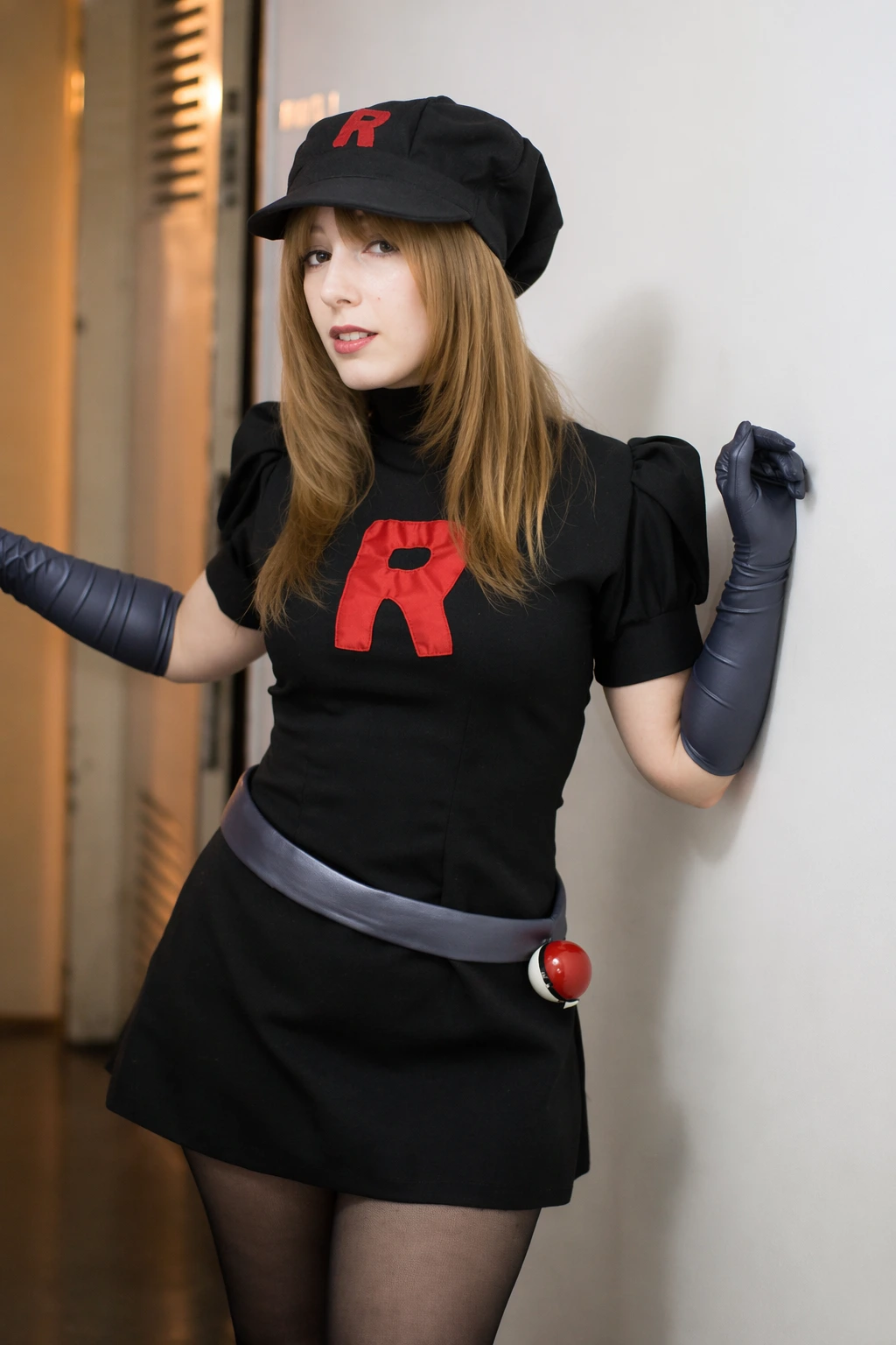 Black mini dress cosplay with red logo, cap, gloves, sheer tights, and a playful villainous stance.