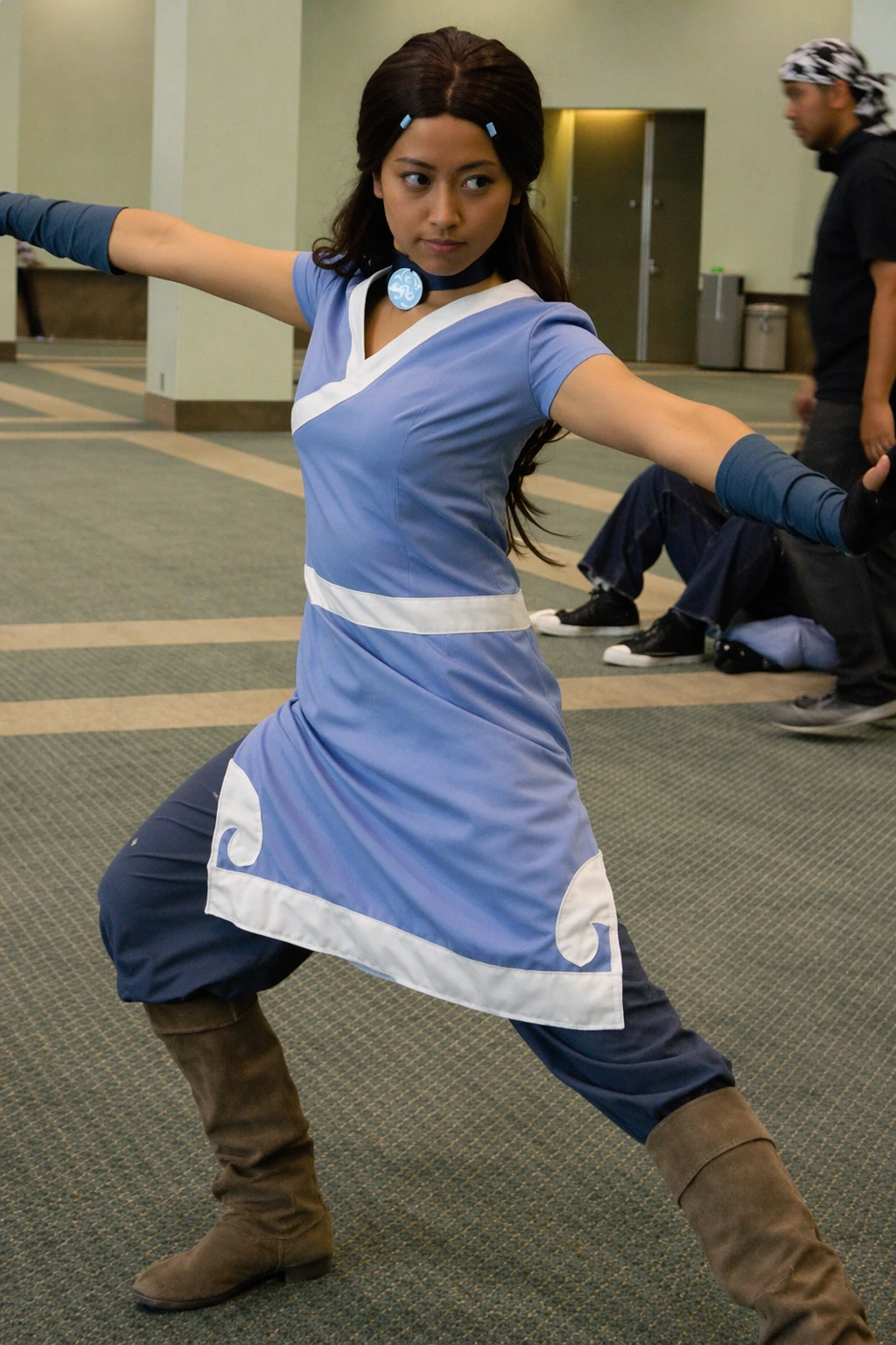 Cosplayer strikes wide martial pose indoors, wearing light blue tunic, dark pants, gloves, and brown boots.