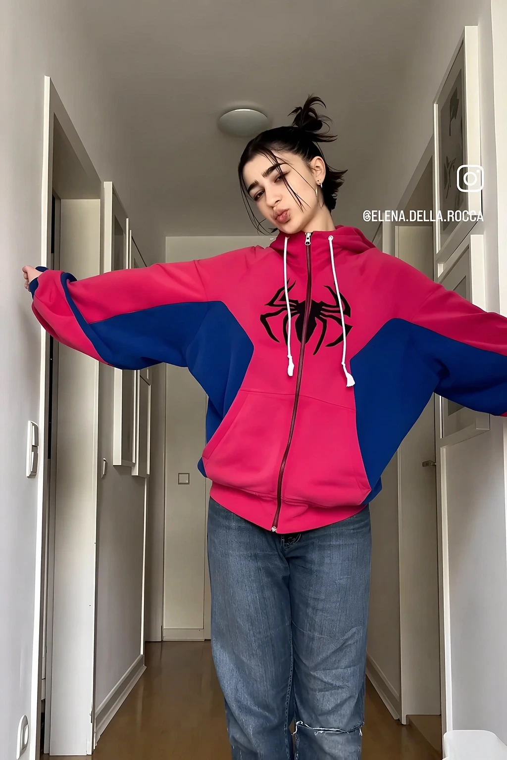 Young woman in oversized pink-and-blue hoodie with black spider logo, loose jeans, and casual hallway pose.