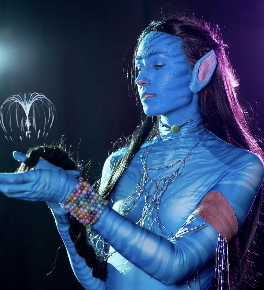 Blue-painted avatar performer in profile, long dark hair, pointed ear prosthetics, layered necklaces, and colorful bead cuff.