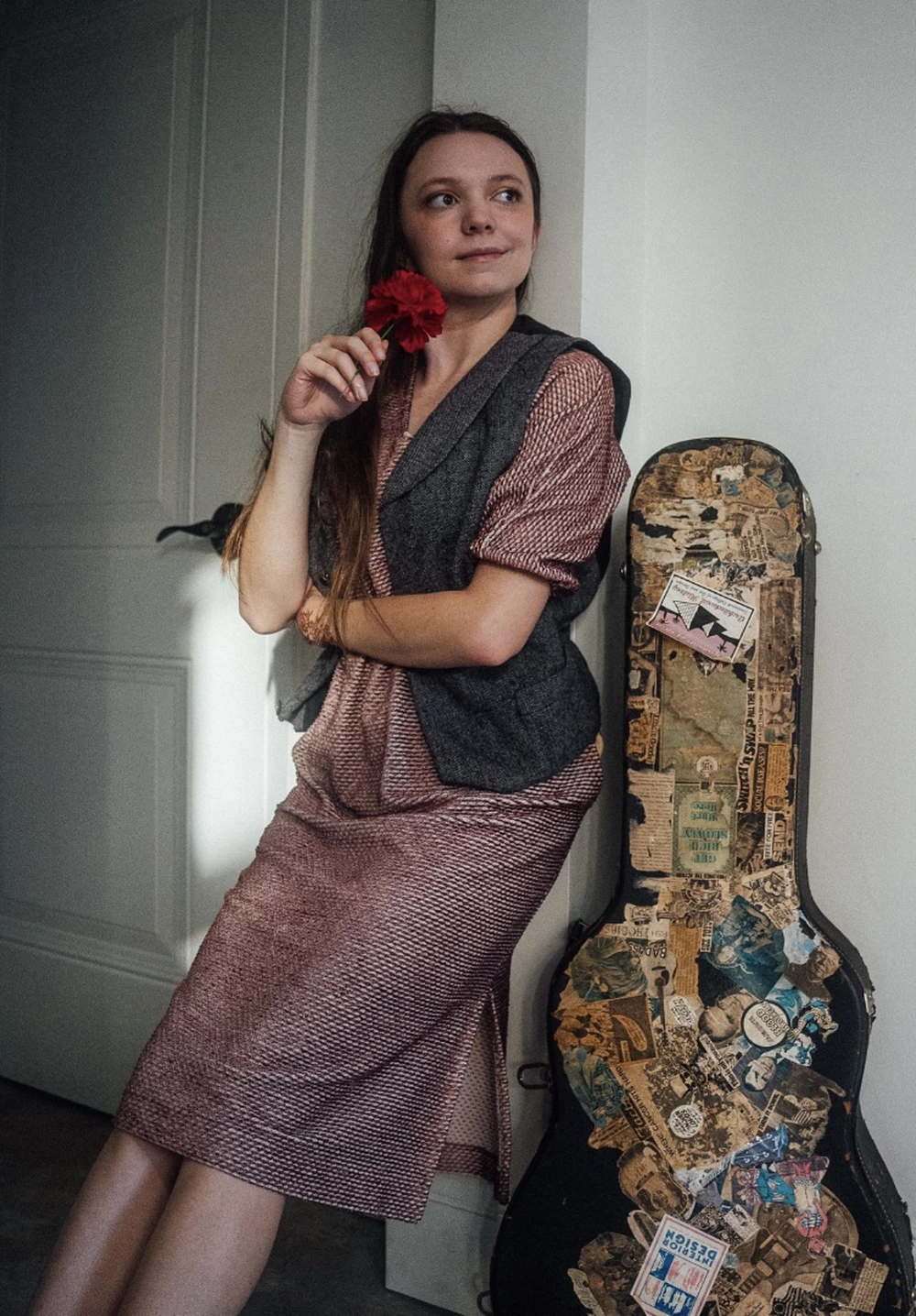 Woman leans indoors in a mauve patterned dress, charcoal vest, holding a red flower beside a sticker-covered guitar case.