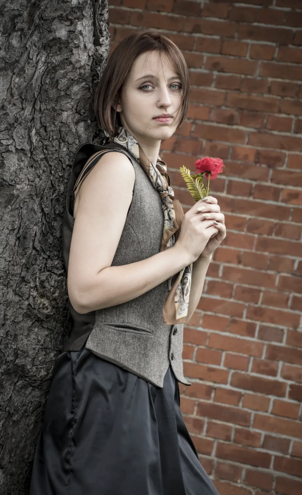 Short bob haircut, gray vest, black skirt, patterned neck scarf, holding a red flower beside a rough tree.