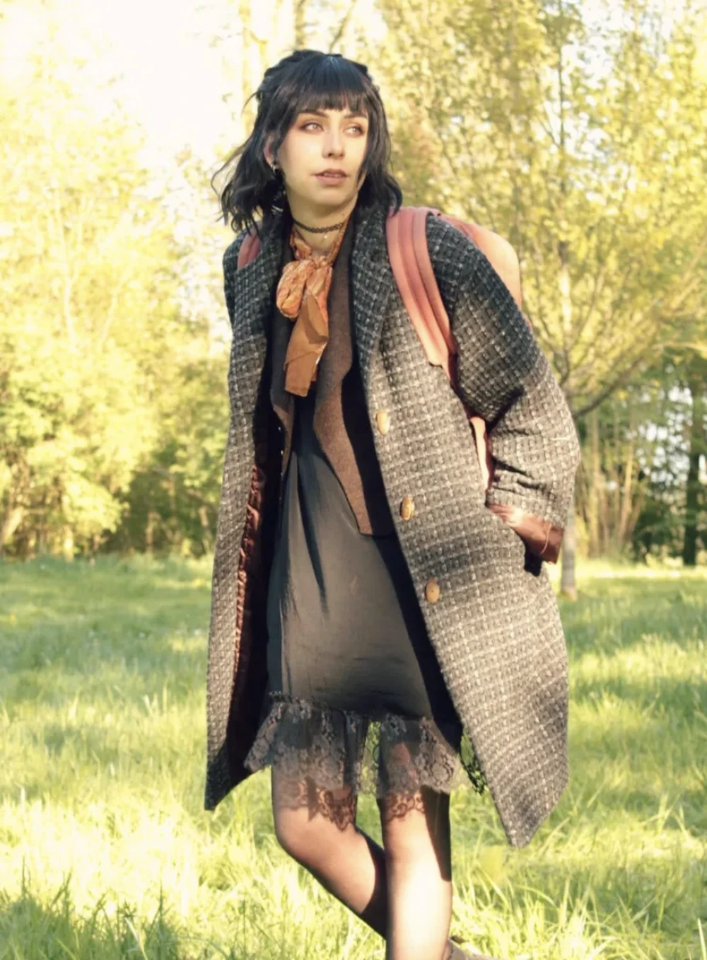 Woman in a dark tweed coat, sheer black dress, peach scarf, and pink backpack walking outdoors.