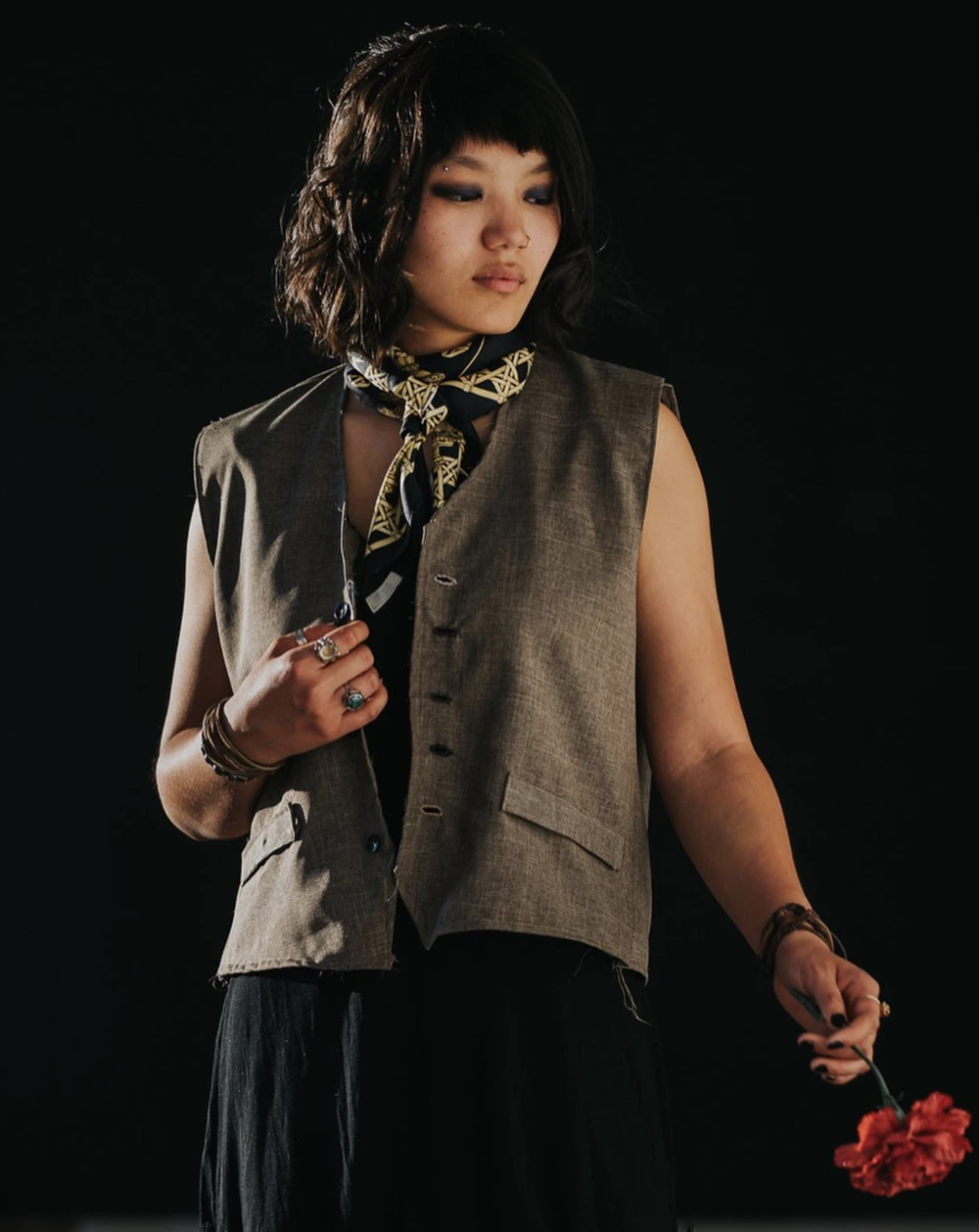 Model wearing a sleeveless brown vest, black outfit, patterned neck scarf, and holding a red flower.