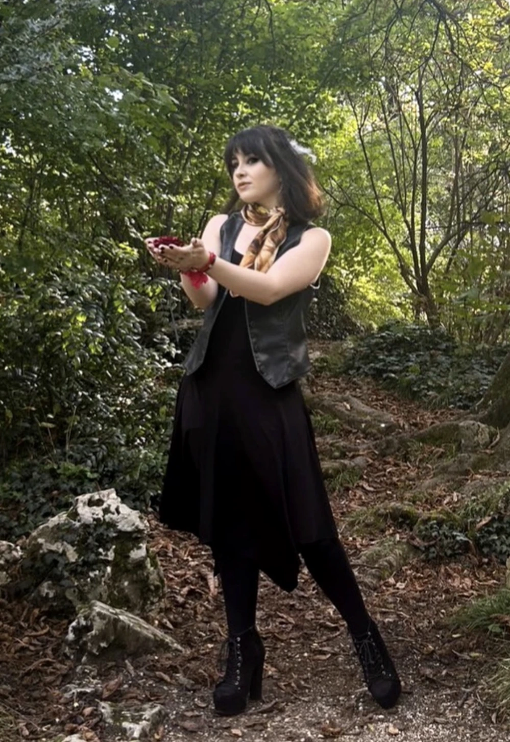 Woman standing on a wooded path wearing a black dress, leather vest, scarf, and heeled lace-up boots.