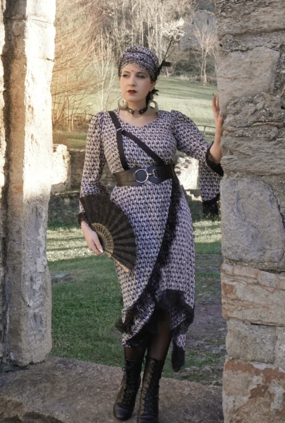 Woman poses by stone ruins wearing a long patterned dress, matching headwrap, belt, boots, and handheld fan.