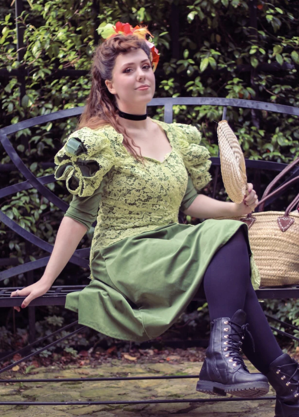 Woman seated on a bench in a light green lace dress, black tights, combat boots, and a floral headpiece.