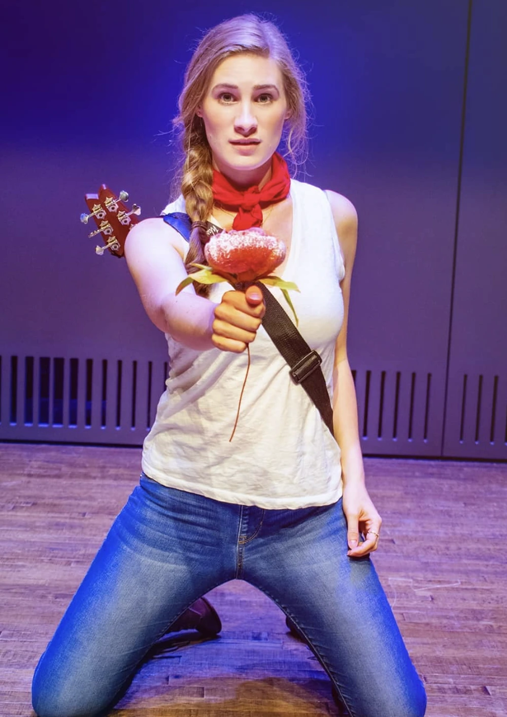 Blonde woman kneels onstage in white tank top, blue jeans, red neck scarf, holding a flower and guitar.