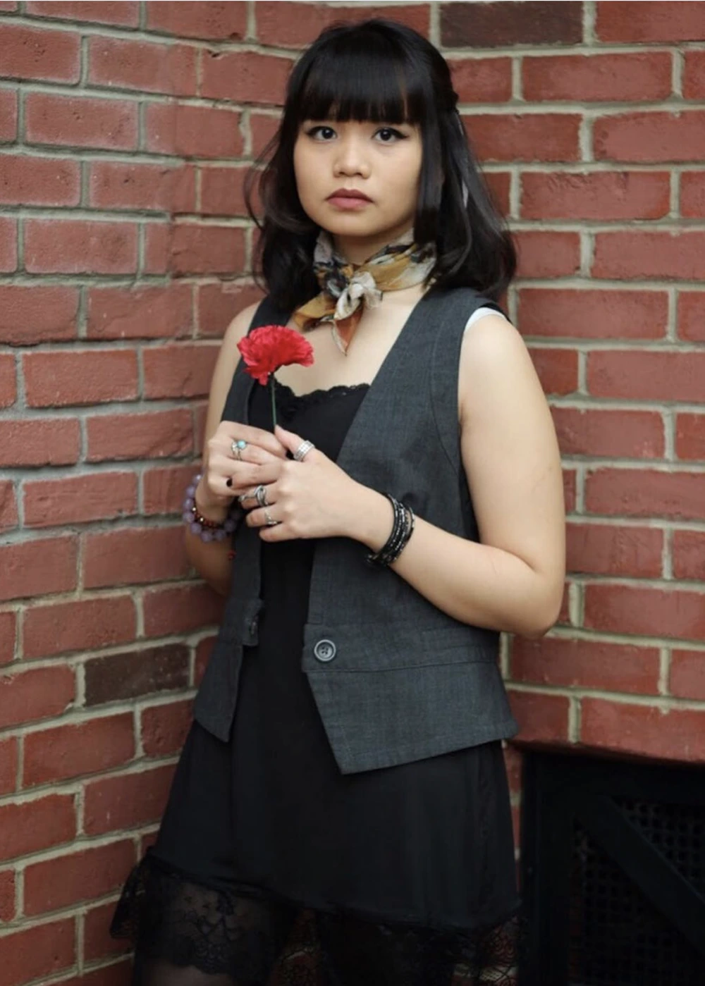Woman in black dress, gray vest, and scarf holds a red carnation against a red brick wall.