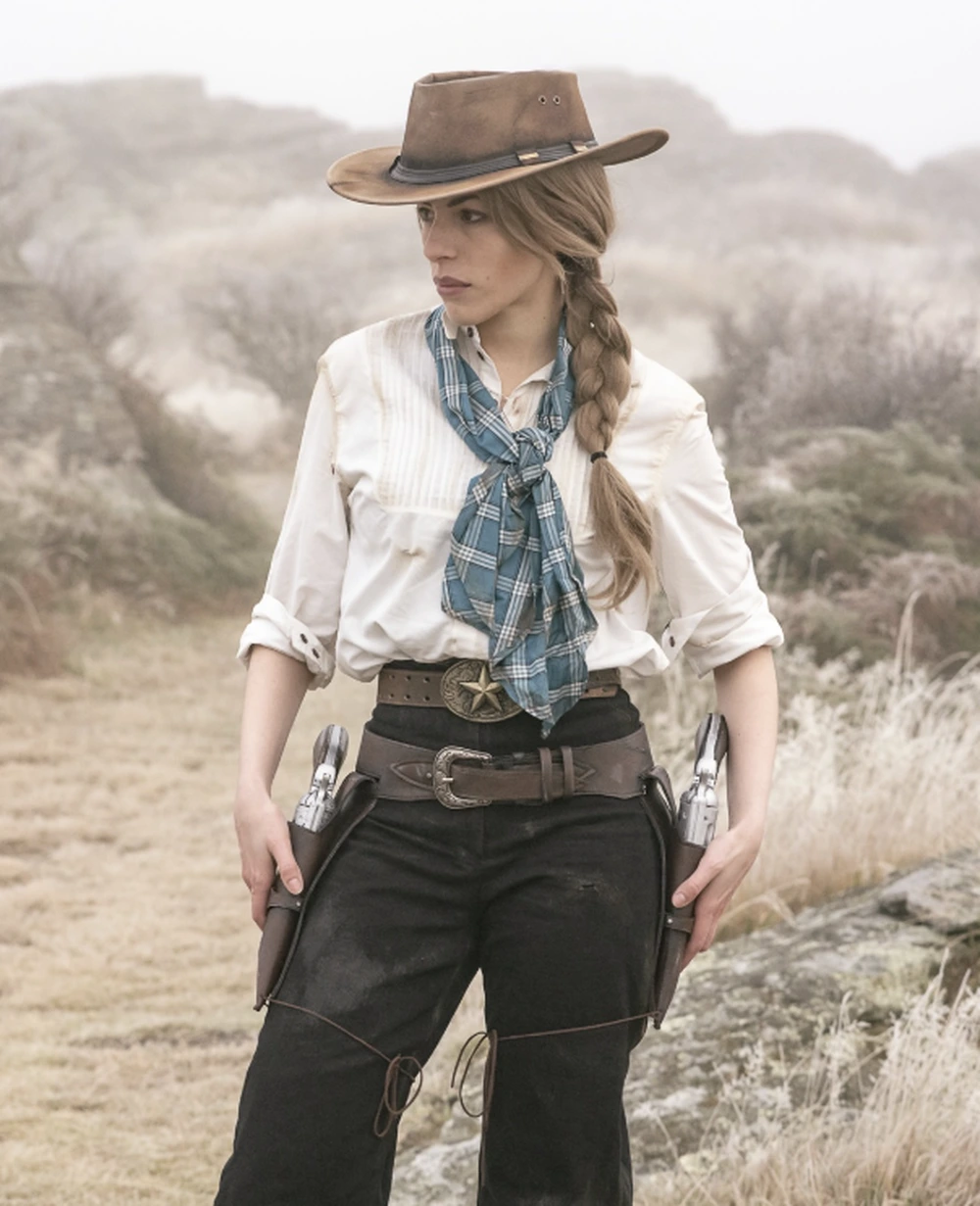 White button-up shirt, blue plaid neckerchief, brown cowboy hat, black pants, twin holsters, standing in scrubby terrain.