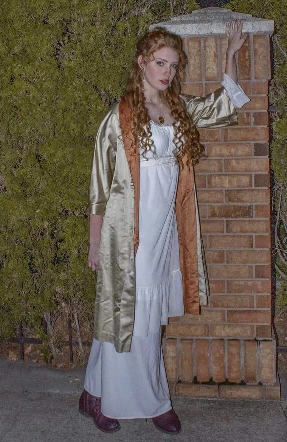 White flowing dress, gold satin robe, long red curls, burgundy shoes, posed outdoors beside a brick pillar.