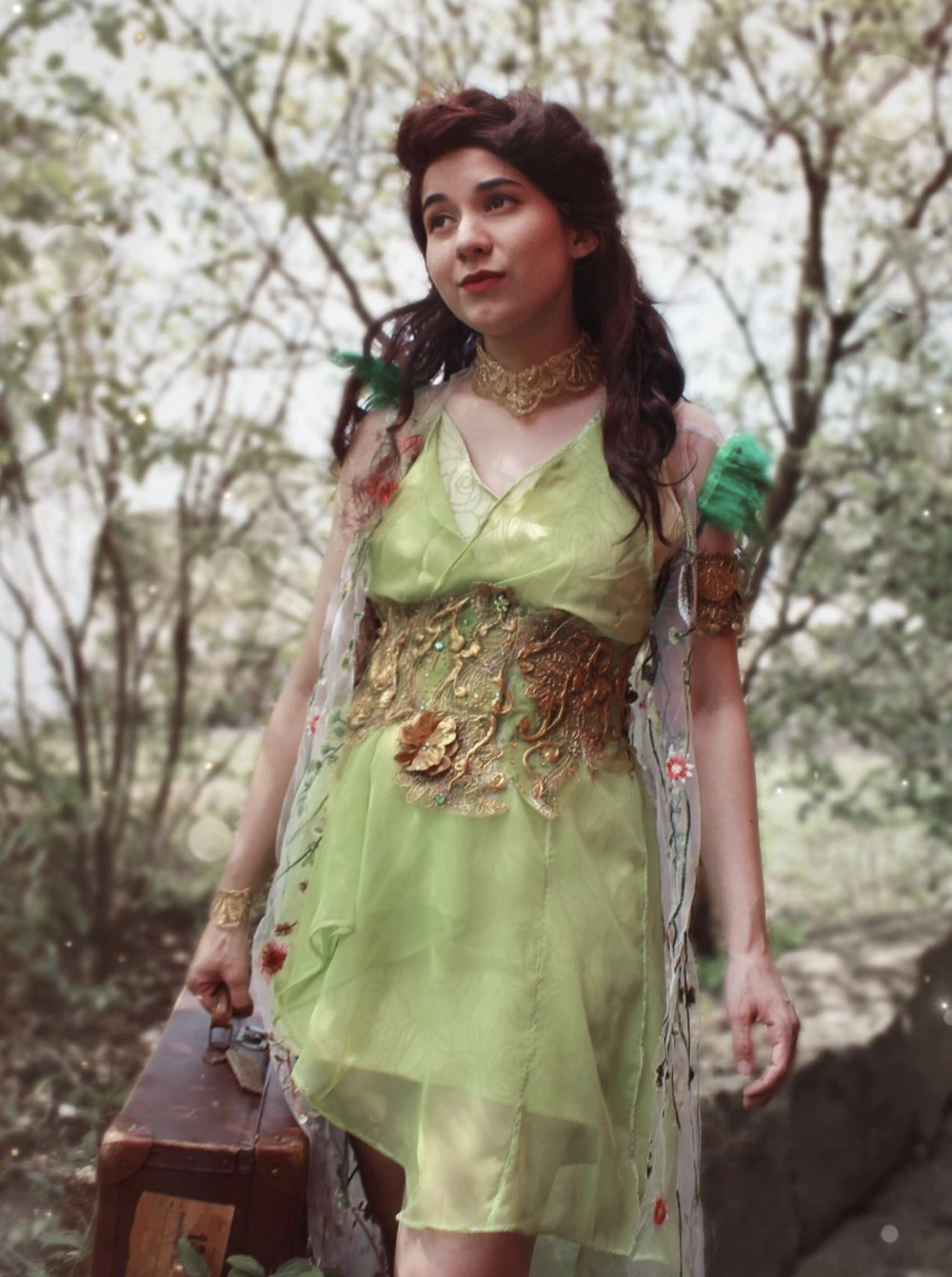 Woman in light green layered dress with gold waist embellishment, floral drape, and suitcase beside her.