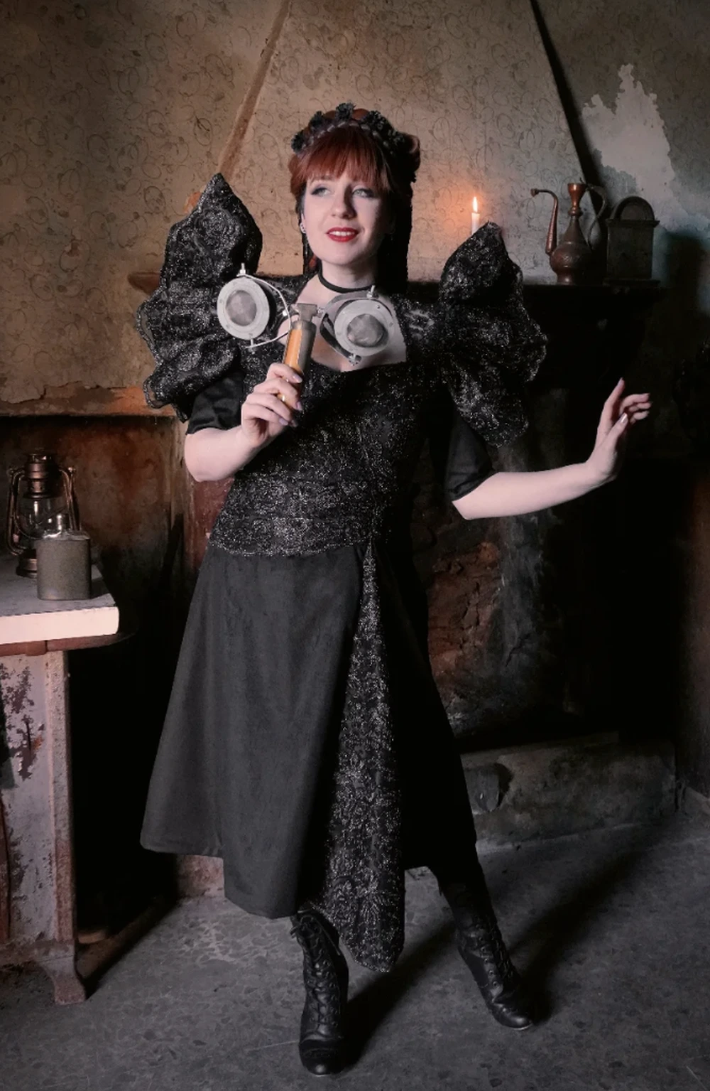 Woman in a dramatic black dress with puffed shoulders, lace details, goggles, and vintage-inspired accessories indoors.