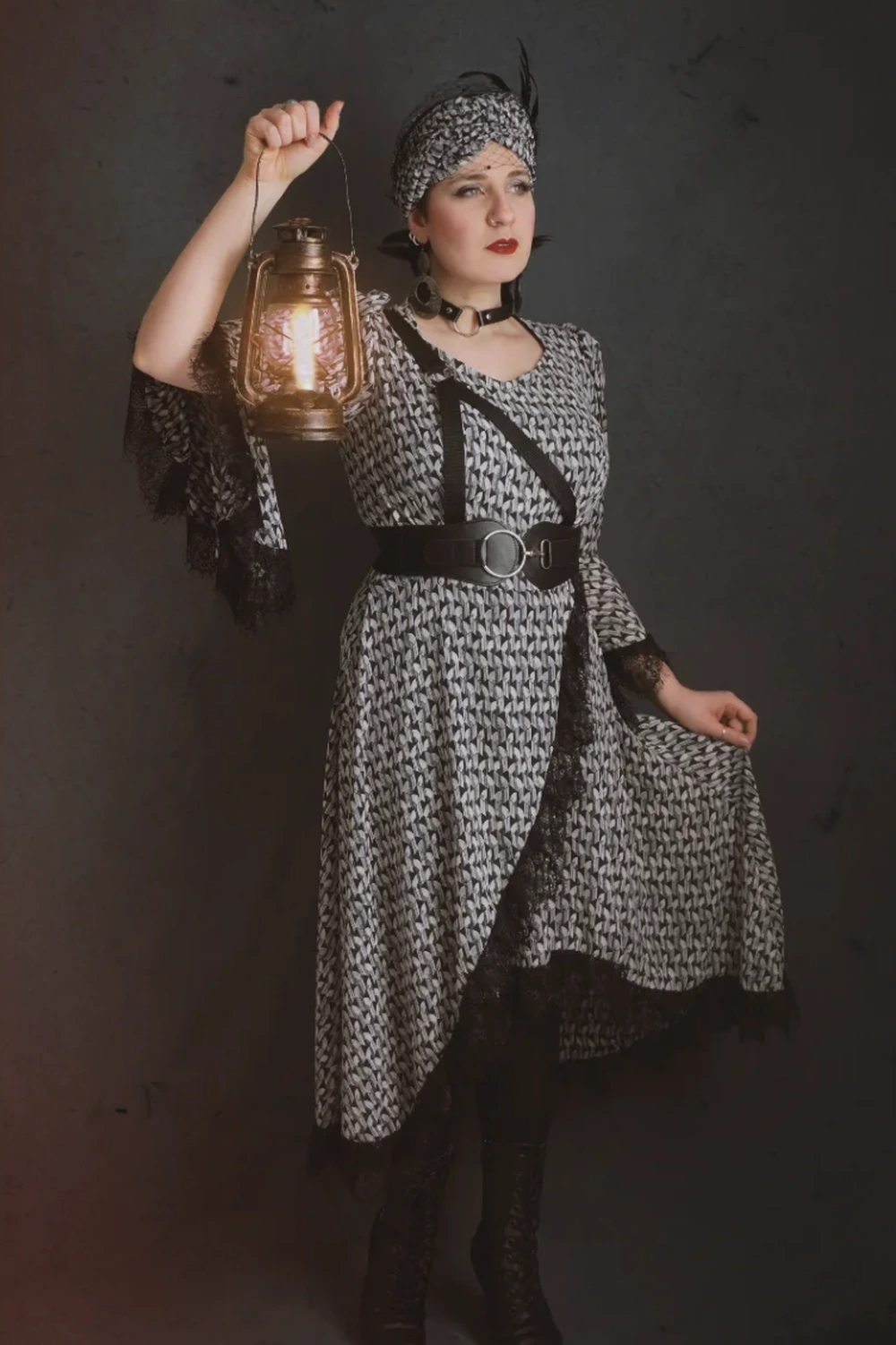 Woman holding a glowing lantern while wearing a patterned black-and-white dress, headwrap, belt, and lace-trimmed sleeves.