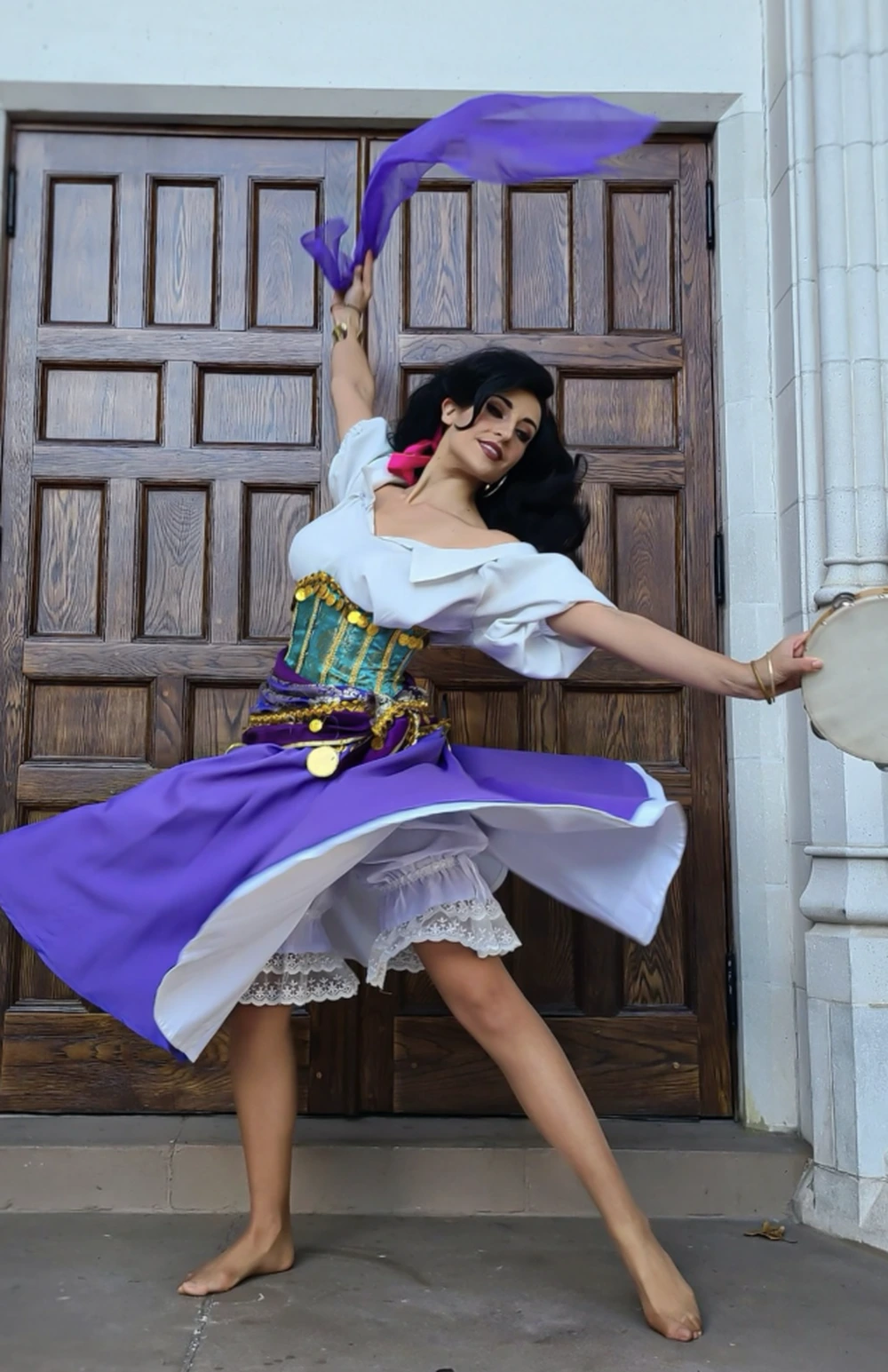 Woman in a purple-and-white swirling dress with corset styling, holding sheer fabric and a tambourine, mid-spin.